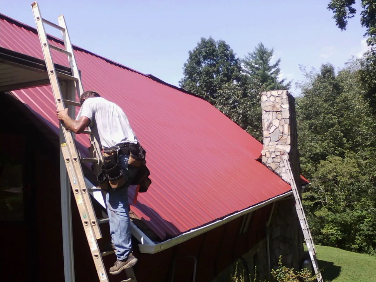 Roofer on a ladder beside a red metal roof and stone chimney — Soffit & Fascia Repair by Roofing Brothers in Kenbridge
